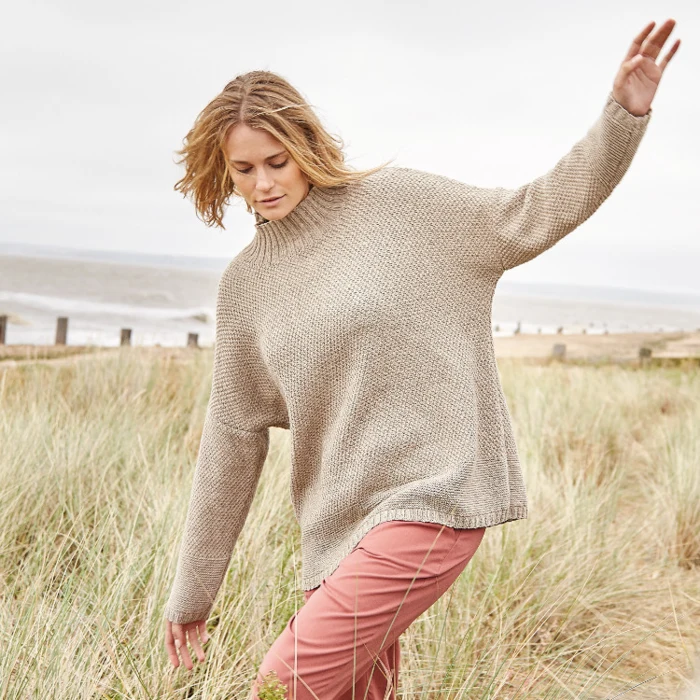 Billede af model iklædt beige Seaham-sweater på en baggrund af et strandlandskab.